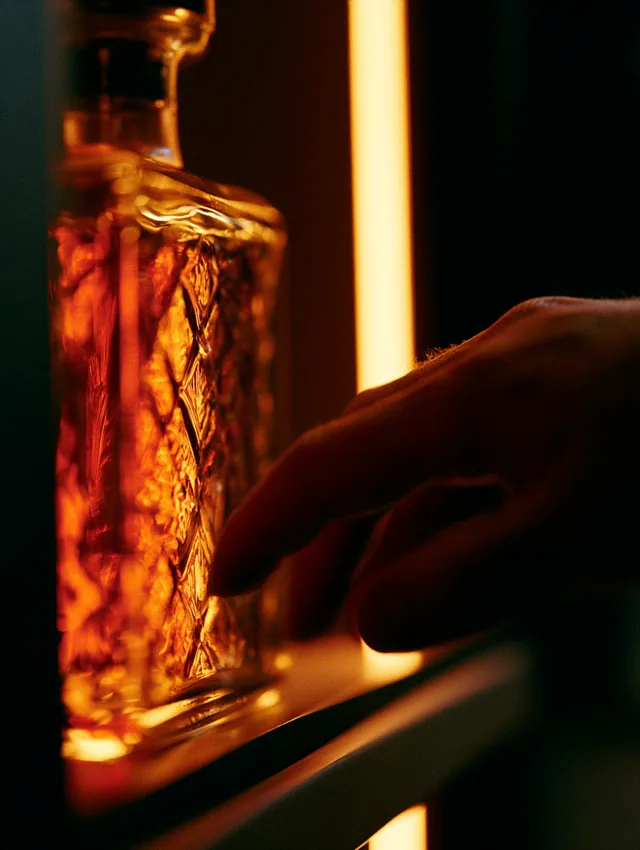 A hand reaching toward a coveted whiskey bottle on a softly lit shelf